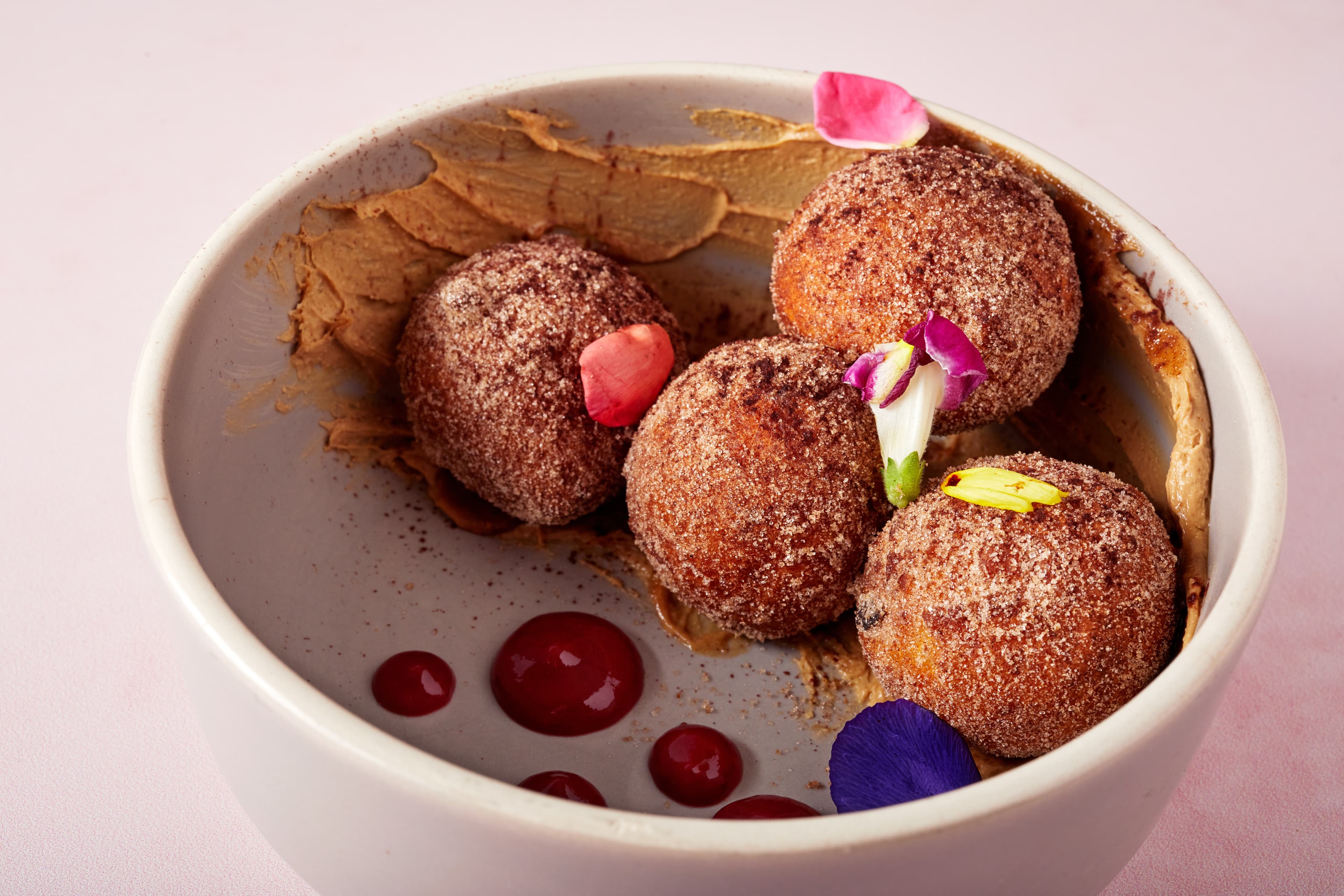 Close-up of cinnamon sugar-dusted zeppole topped with edible flower petals, served in a bowl with espresso mascarpone, house jam, and decorative raspberry sauce on a pink background.