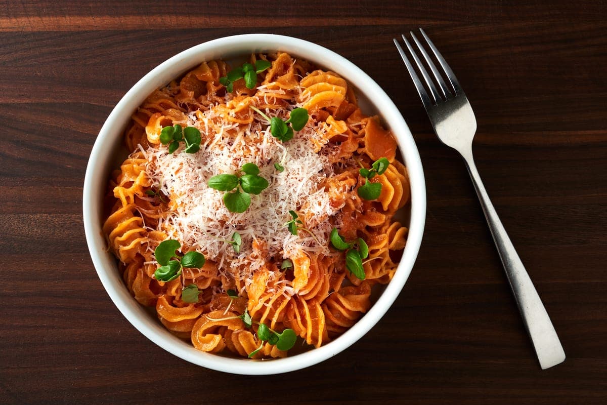 Overhead view of a bowl of radiatore pasta tossed in creamy vodka sauce, topped with grated Parmesan cheese and fresh microgreens, served on a dark wood table with a fork beside the bowl.