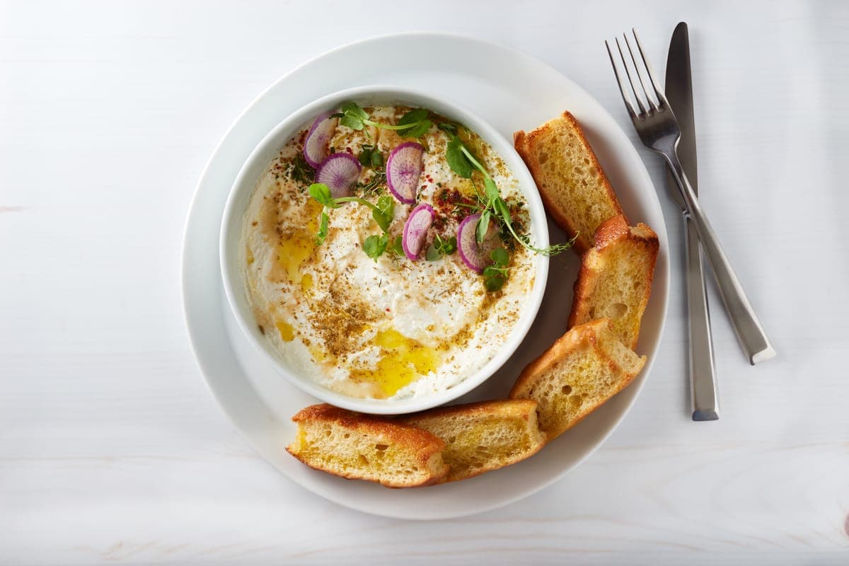 A bowl of housemade whipped ricotta drizzled with olive oil and garnished with thinly sliced radishes, herbs, and spices, served with toasted slices of golden focaccia on a white plate with fork and knife.