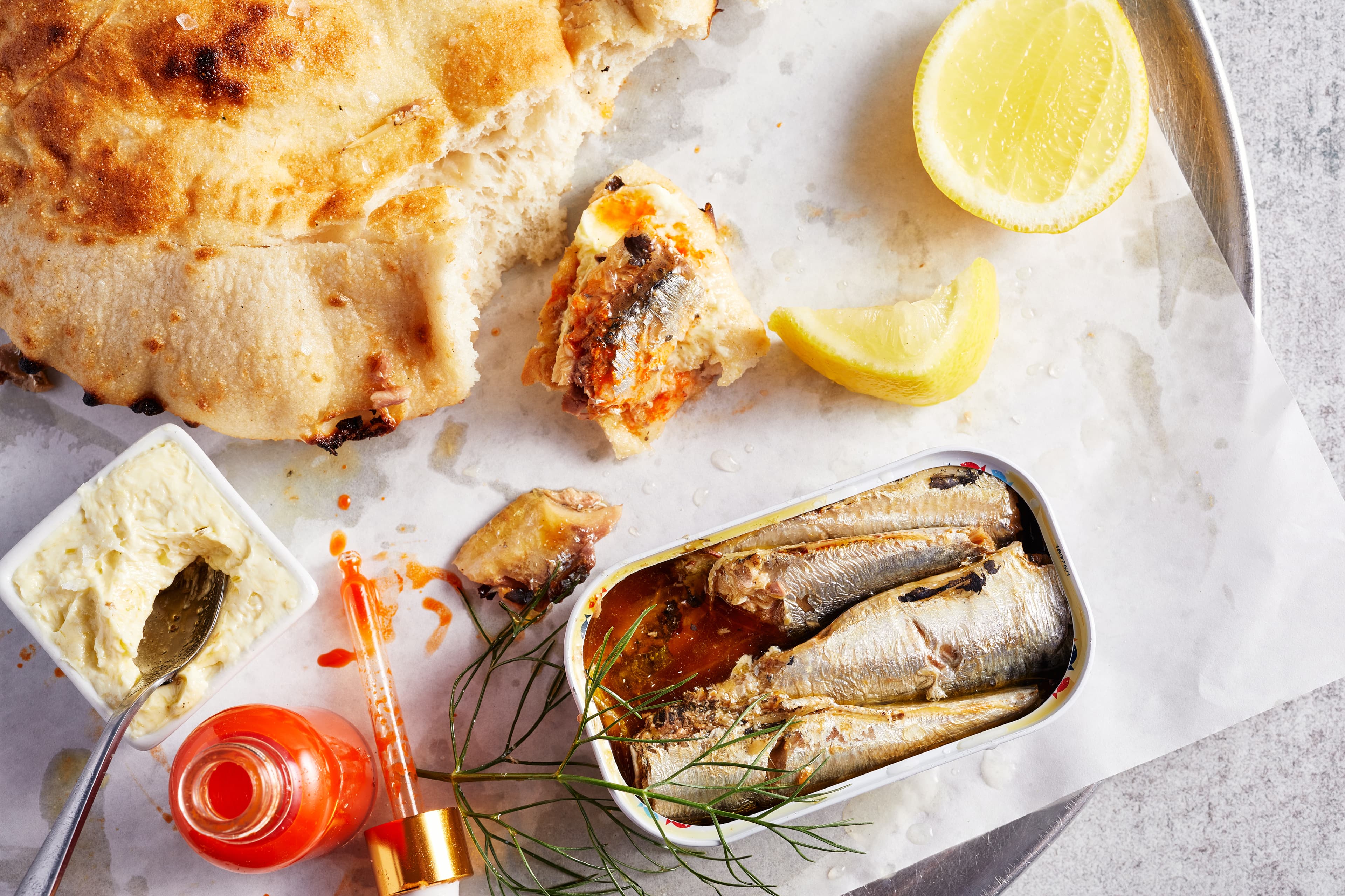 Overhead view of Sofia’s Sardines antipasto featuring garlic butter-toasted piada bread, tinned sardines in chili oil, lemon wedges, Fresno hot sauce with dropper, and whipped butter on parchment.
